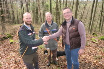 Drei Männer stehen im Wald. Zwei schütteln sich die Hände.